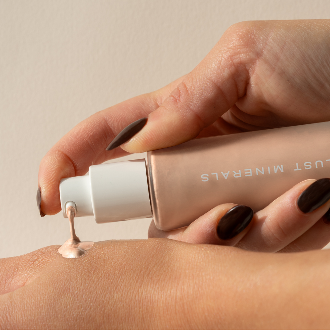 Hand applying liquid Illuminator  from a tube labeled 'Just Minerals' on a beige background