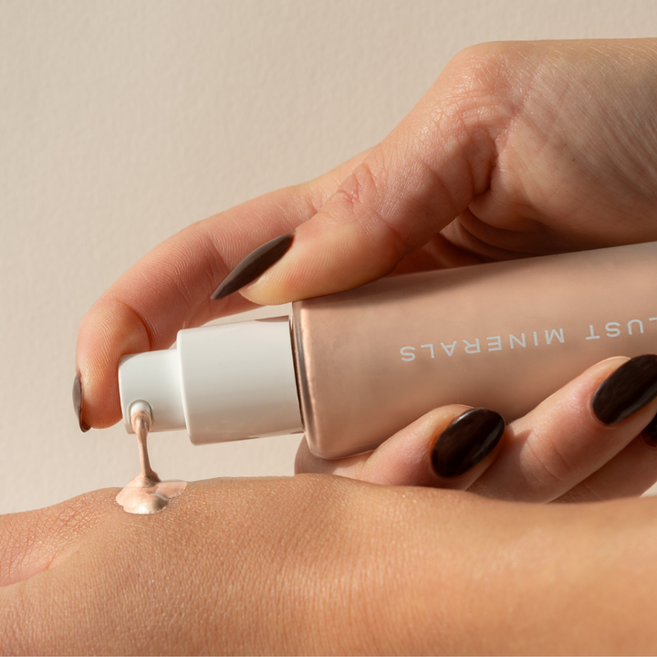 Hand applying liquid Illuminator  from a tube labeled 'Just Minerals' on a beige background