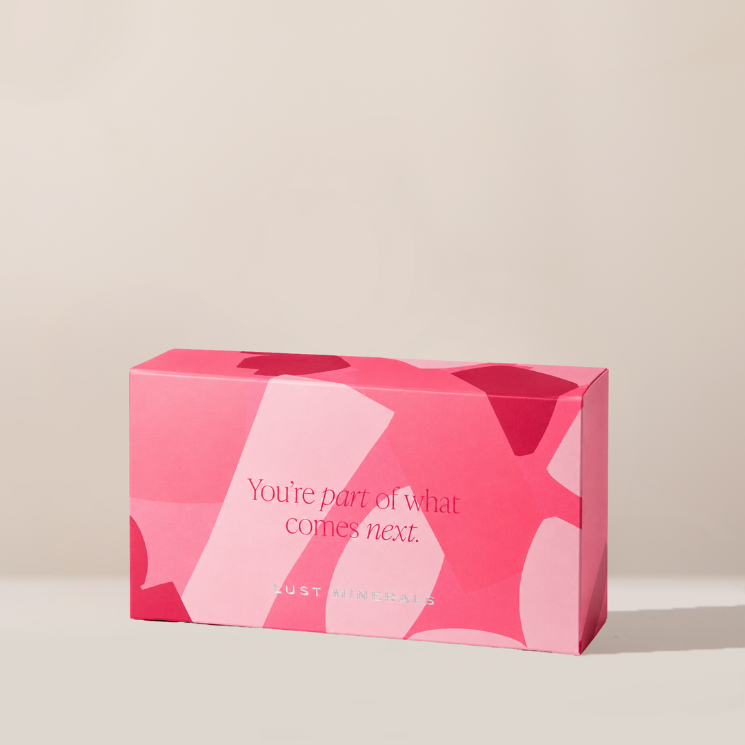 A pink rectangular product box with abstract geometric shapes and the text 'You're part of what comes next.' and 'LUST MINERALS' printed on the front, photographed on a clean warm beige background.